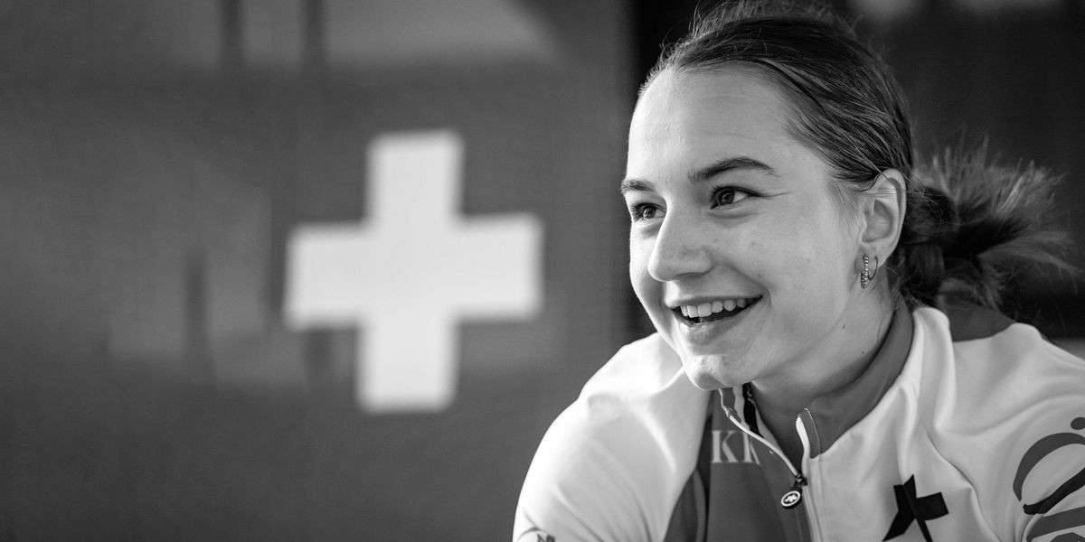 Jeune femme souriante portant une tenue de cyclisme, avec un drapeau suisse en arrière-plan.