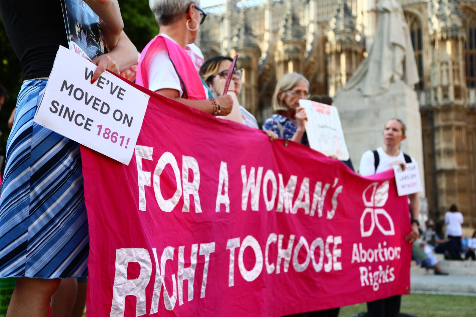 Gruppe von Menschen hält ein grosses rosa Banner mit der Aufschrift ’For a woman’s right to choose – Abortion Rights’ vor einem historischen Gebäude. Eine Person hält ein Schild mit der Aufschrift ’We’ve moved on since 1861’.