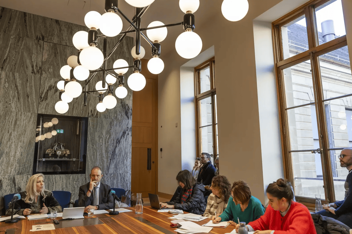 Réunion d’affaires dans une salle moderne avec un grand lustre et des fenêtres, plusieurs personnes travaillent autour d’une table.