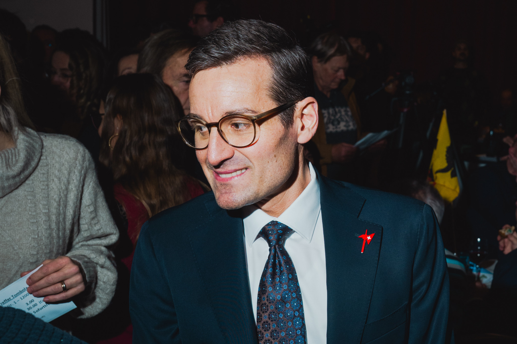 Mann in Anzug mit Brille und roter Anstecknadel lächelt in einer Menschenmenge.