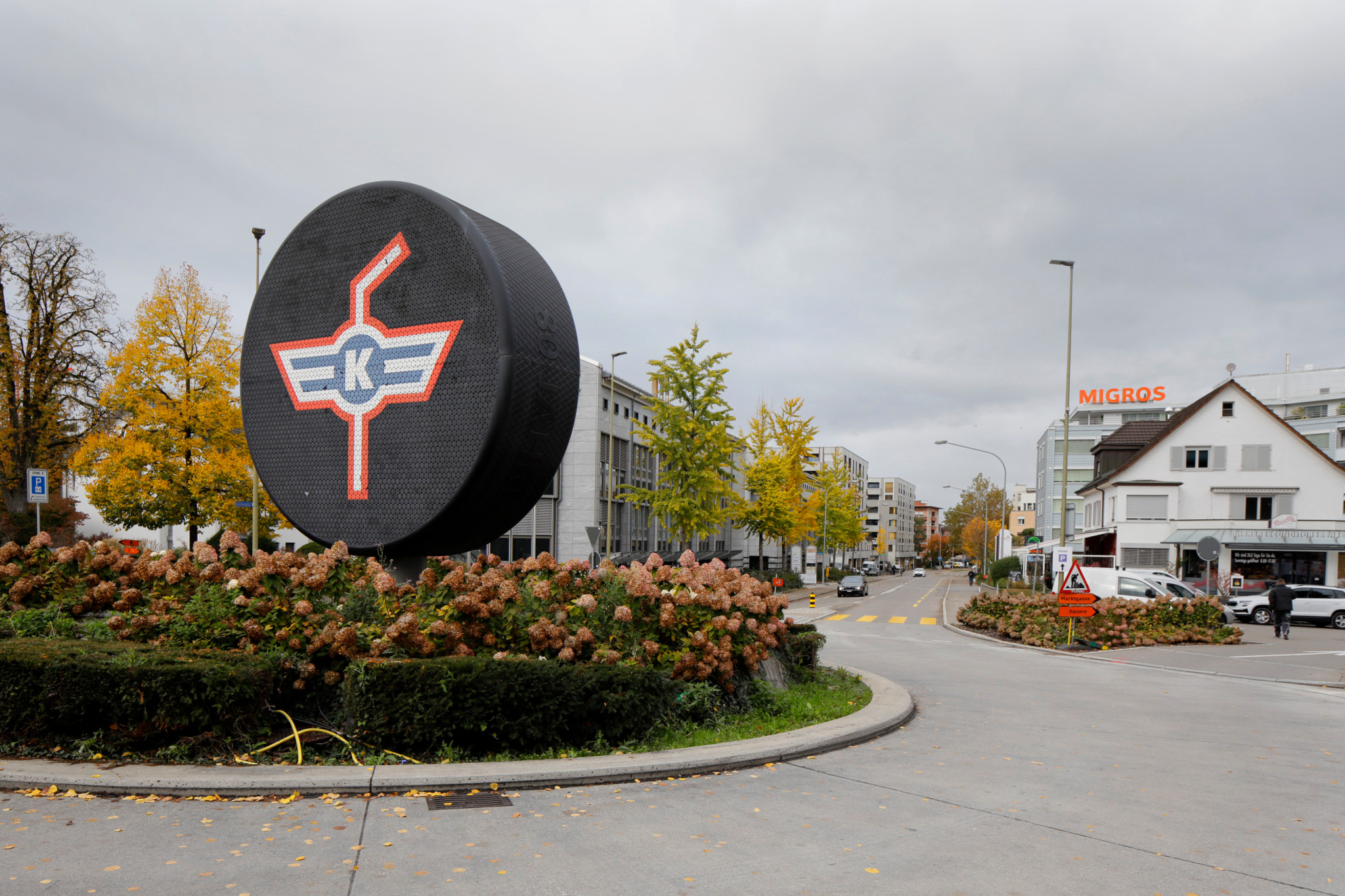 Grosser schwarzer Puck mit K-Logo auf einem Kreisel an der Schaffhauserstrasse in Kloten, mit Häusern und herbstlichen Bäumen im Hintergrund.
