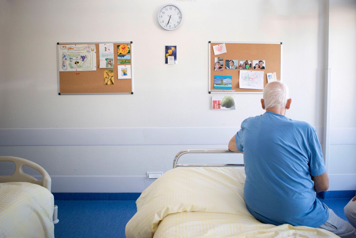 Patient assis sur un lit à l’hôpital de Beaumont, Lausanne, avec des tableaux d’affichage et une horloge murale visibles.