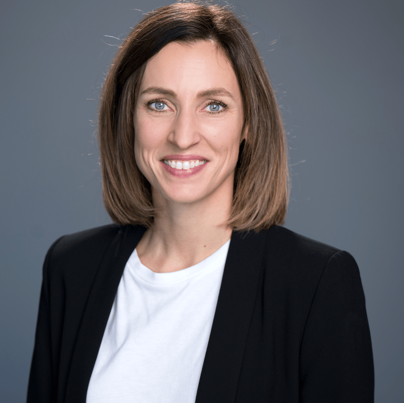 Portrait d’une femme souriante aux cheveux bruns, portant un blazer noir et un t-shirt blanc, sur un fond gris.