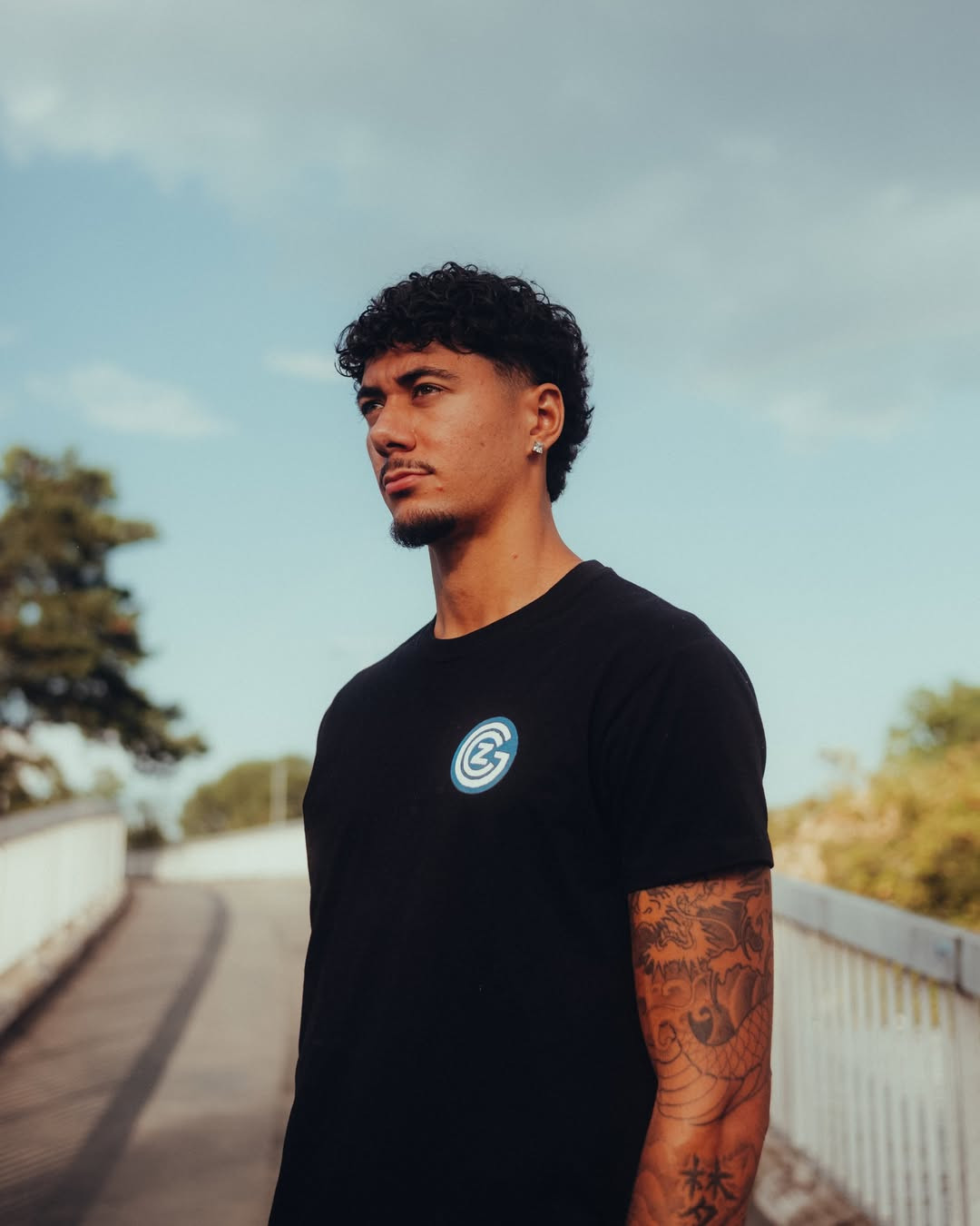 Ein junger Mann mit lockigem Haar steht nachdenklich auf einer Brücke. Er trägt ein schwarzes T-Shirt mit einem blauen Logo und hat Tätowierungen am Arm. Im Hintergrund sind Bäume und ein bewölkter Himmel zu sehen.