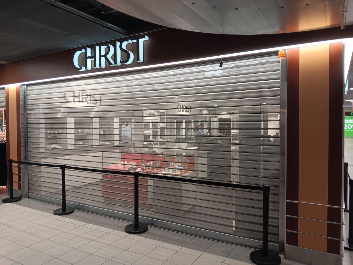Magasin de bijoux fermé avec rideau métallique baissé, enseigne 'Christ' lumineuse au-dessus de la vitrine.