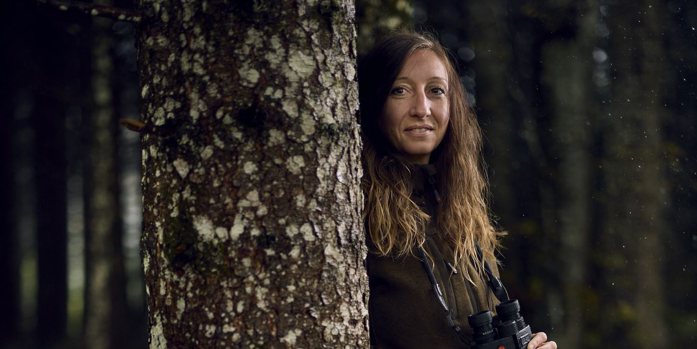 Portrait d'une femme dans une forêt, portant des vêtements de chasse et tenant des jumelles.