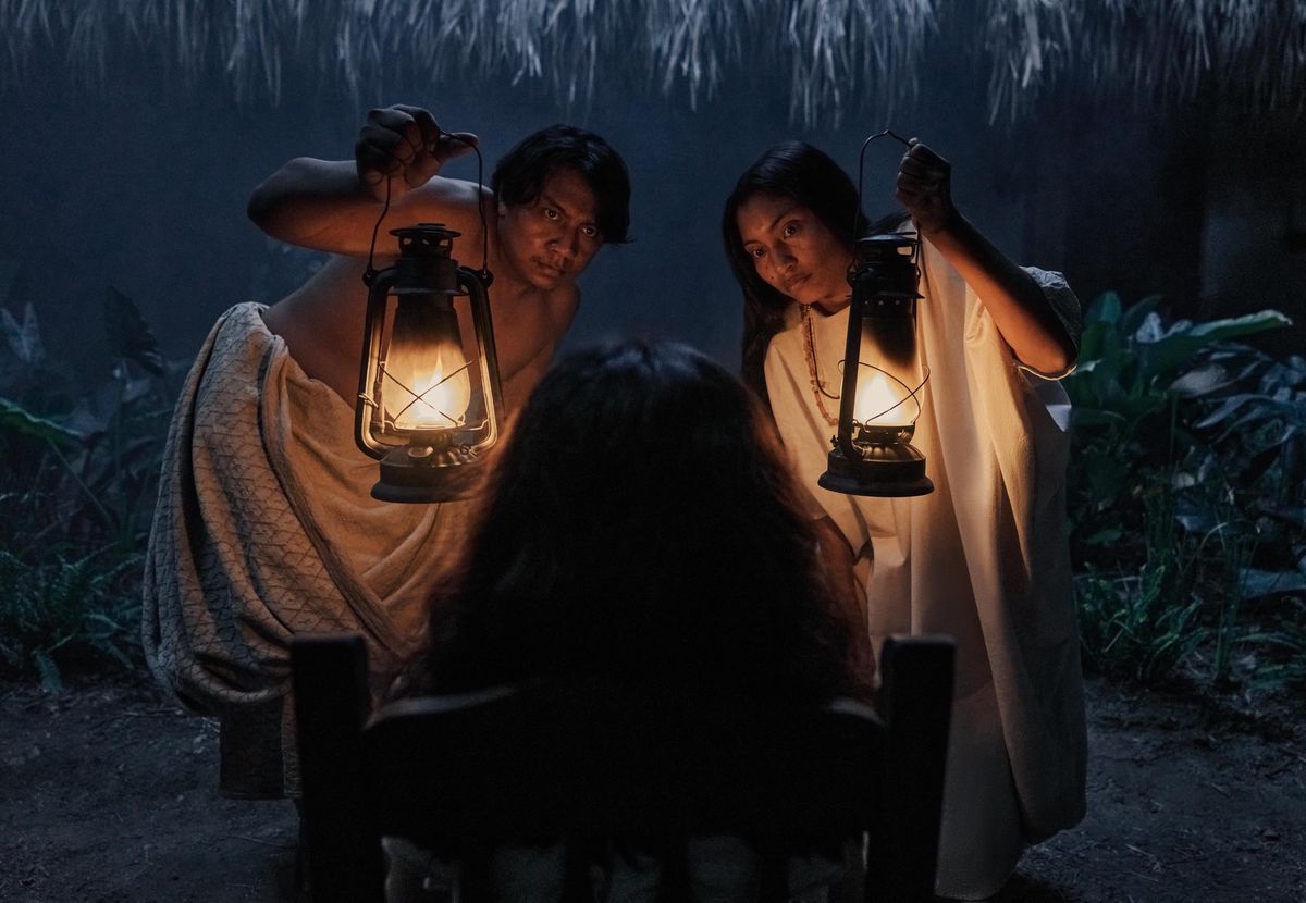Scène sombre avec Andris Pana et Nicole Montenegro tenant des lampes à huile, face à une femme de dos, dans ’Cien Años de Soledad’ S1.