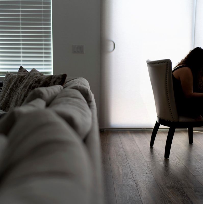 Frau sitzt mit einem Smartphone in einem dunklen Raum, neben einem Sofa. Licht fällt durch ein Fenster mit Jalousien ein.