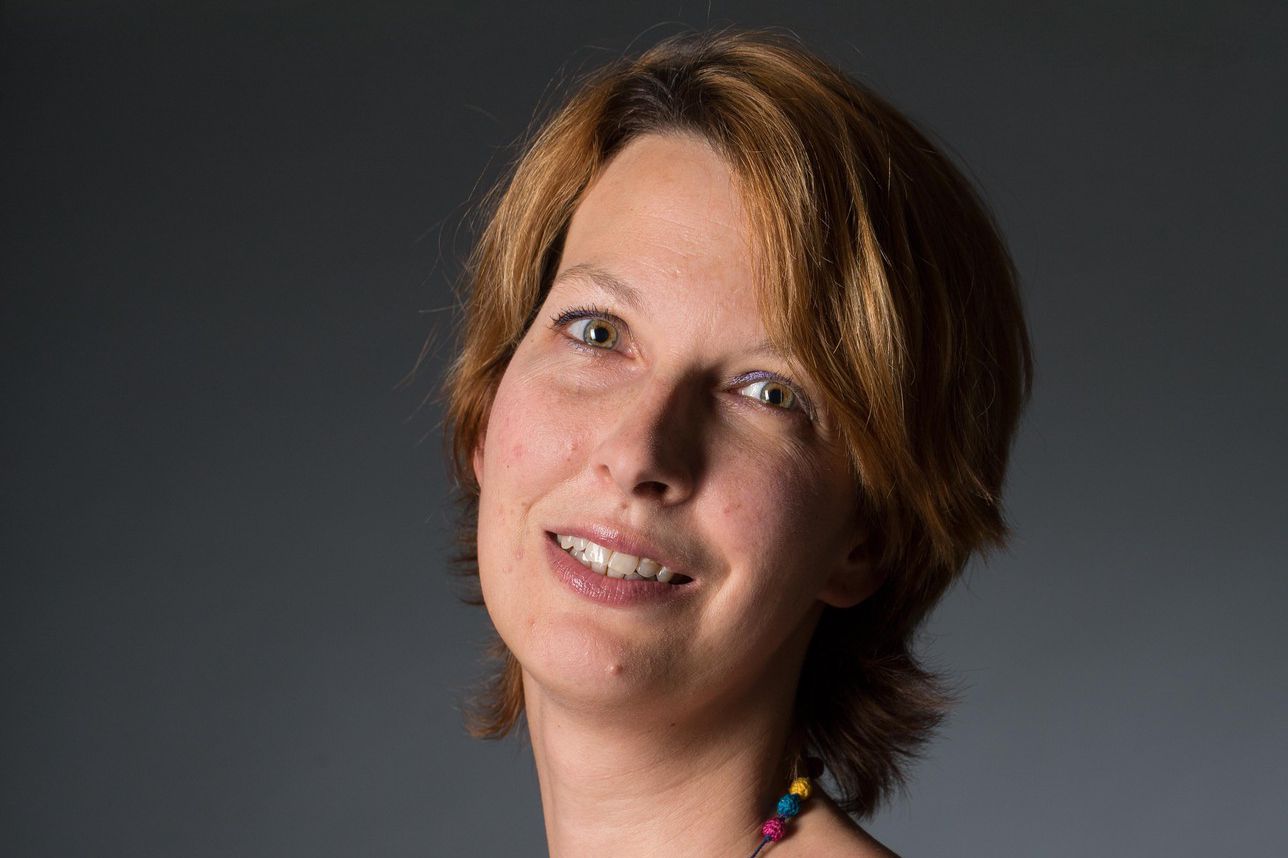 Femme souriante portant un collier coloré et un débardeur gris, posant devant un fond gris.