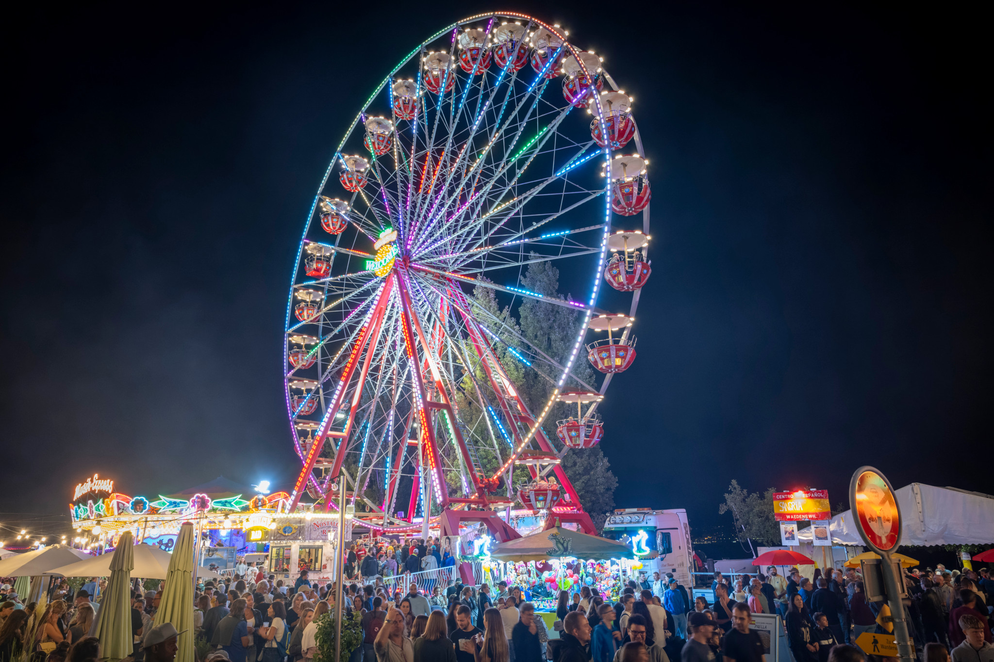 Bunt beleuchtetes Riesenrad bei der Chilbi Wädenswil 2023 bei Nacht, umgeben von einer grossen Menschenmenge und Marktständen.