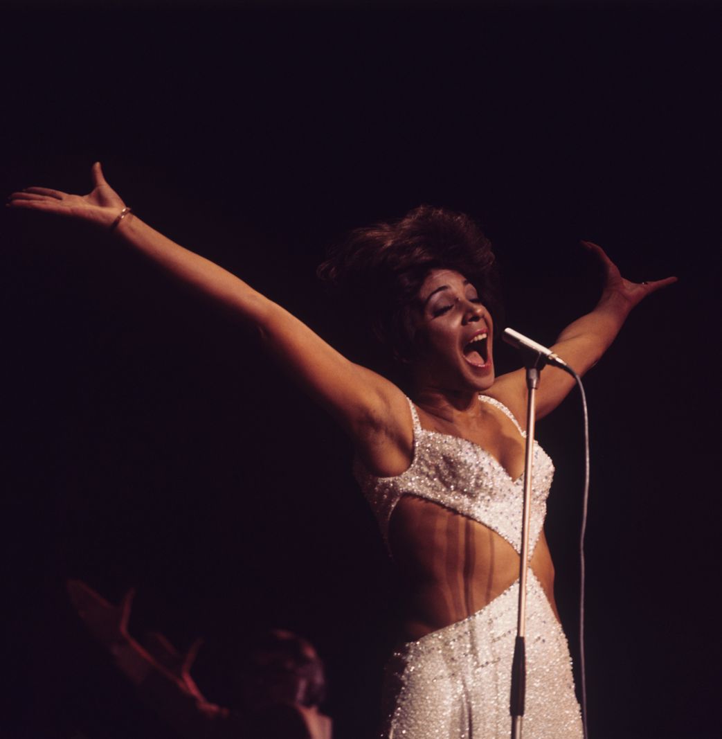 Shirley Bassey sur scène au Royal Variety Performance à Londres, portant une robe blanche scintillante et chantant avec les bras écartés.