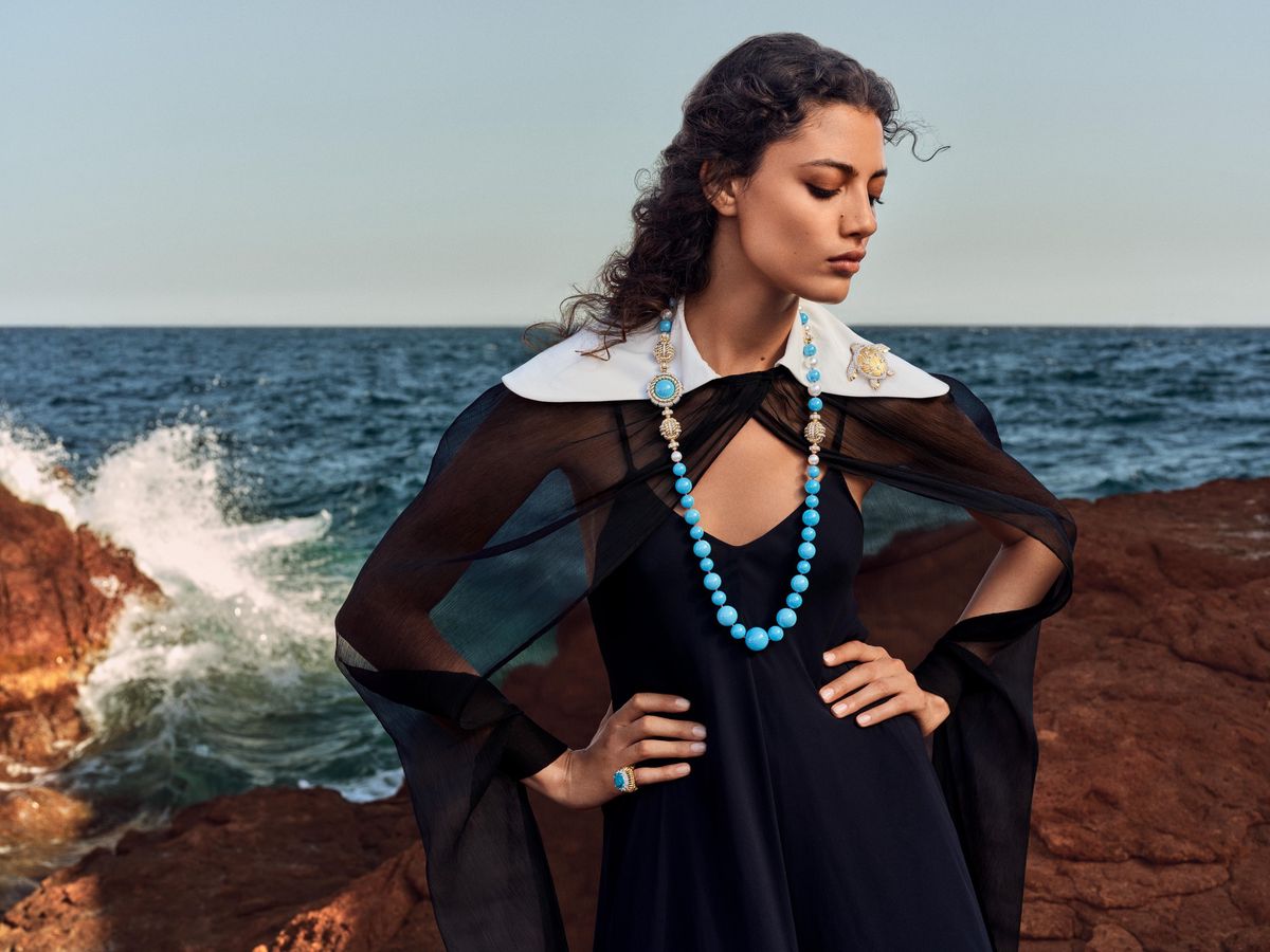 Femme en robe noire et cape transparente, portant un collier bleu, posant près de la mer avec des vagues. Fond de rochers et d’océan.