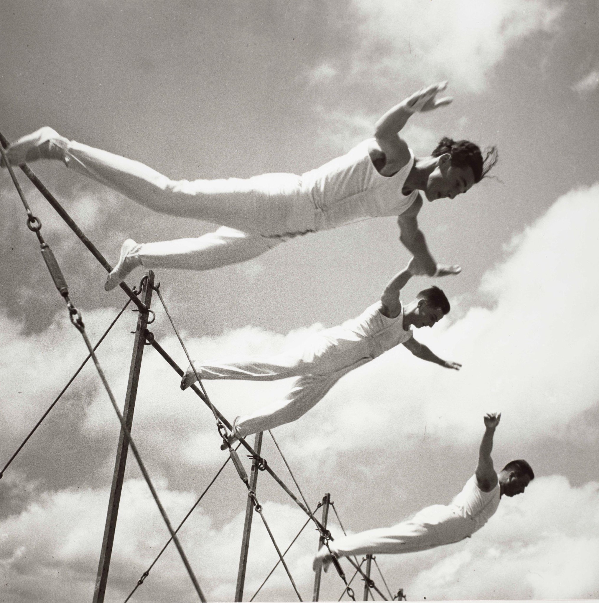 Trois gymnastes exécutant un saut synchronisé sur une barre horizontale sous un ciel nuageux.