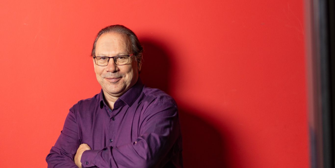 Portrait d'Yves Defferrard, nouveau secrétaire d'Unia Genève, devant une paroi rouge avec le logo Unia écrit en blanc.