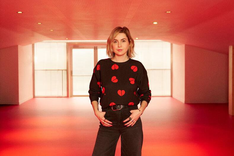 Chloé Thibaud se tenant debout dans une pièce à l’éclairage rouge, portant un pull noir avec des motifs de cœurs rouges.