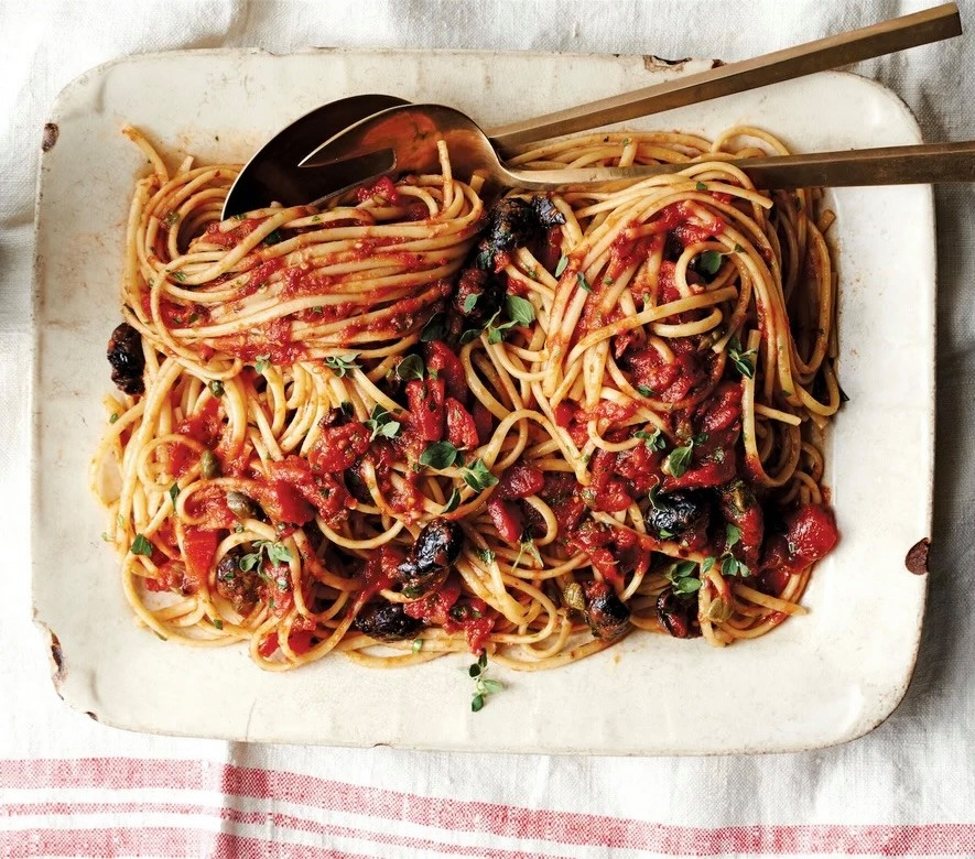 Spaghetti alla Puttanesca mit Tomatensauce, schwarzen Oliven und frischen Kräutern auf einer rustikalen weissen Servierplatte.