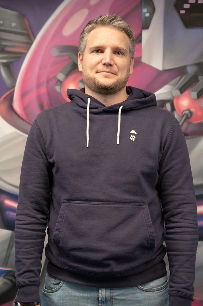 Un homme souriant portant un sweat à capuche bleu foncé se tient devant une œuvre d’art colorée et abstraite.