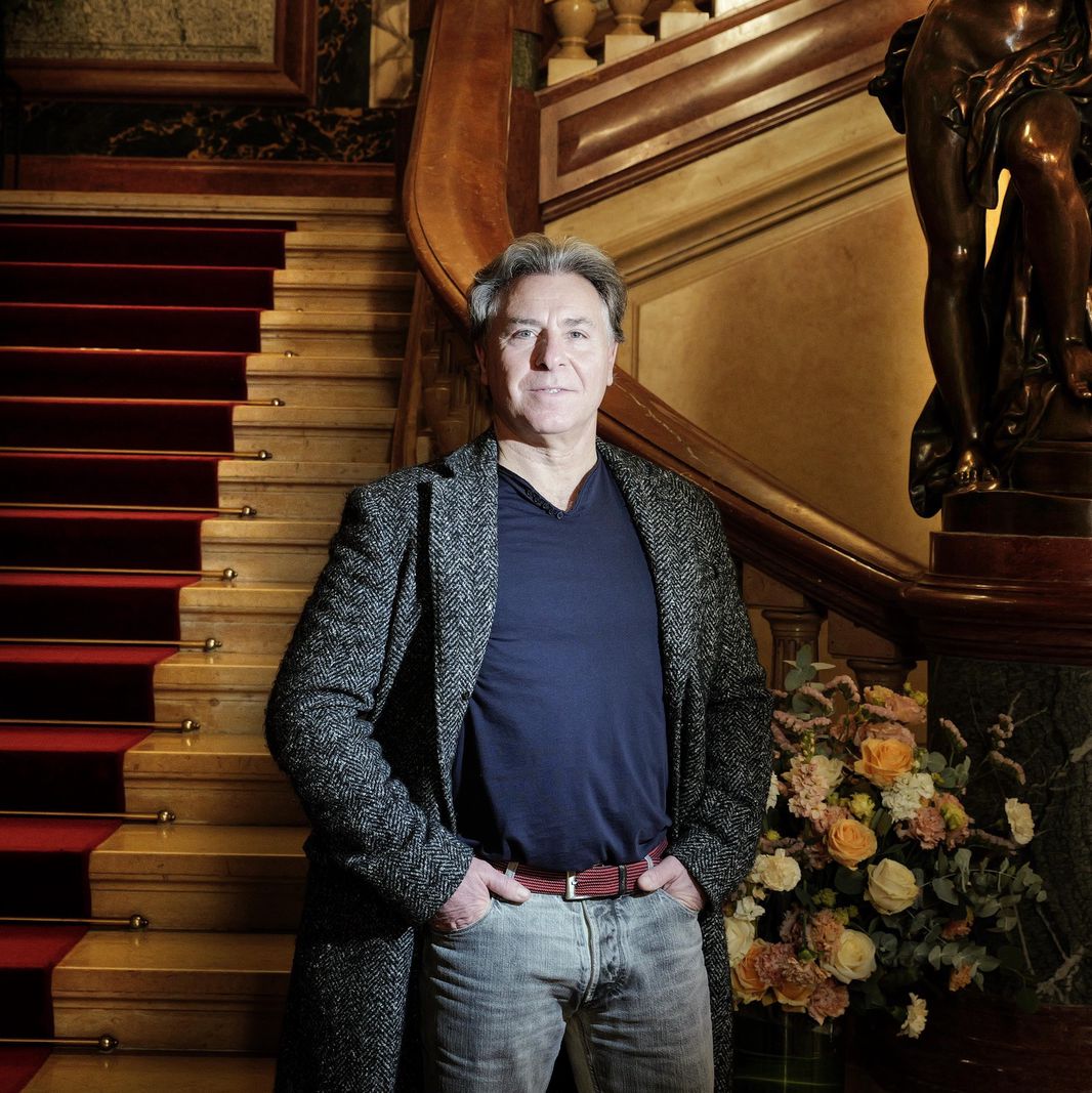 Ténor debout devant un escalier au Grand théâtre de Genève, décor élégant avec statue et bouquet de fleurs.