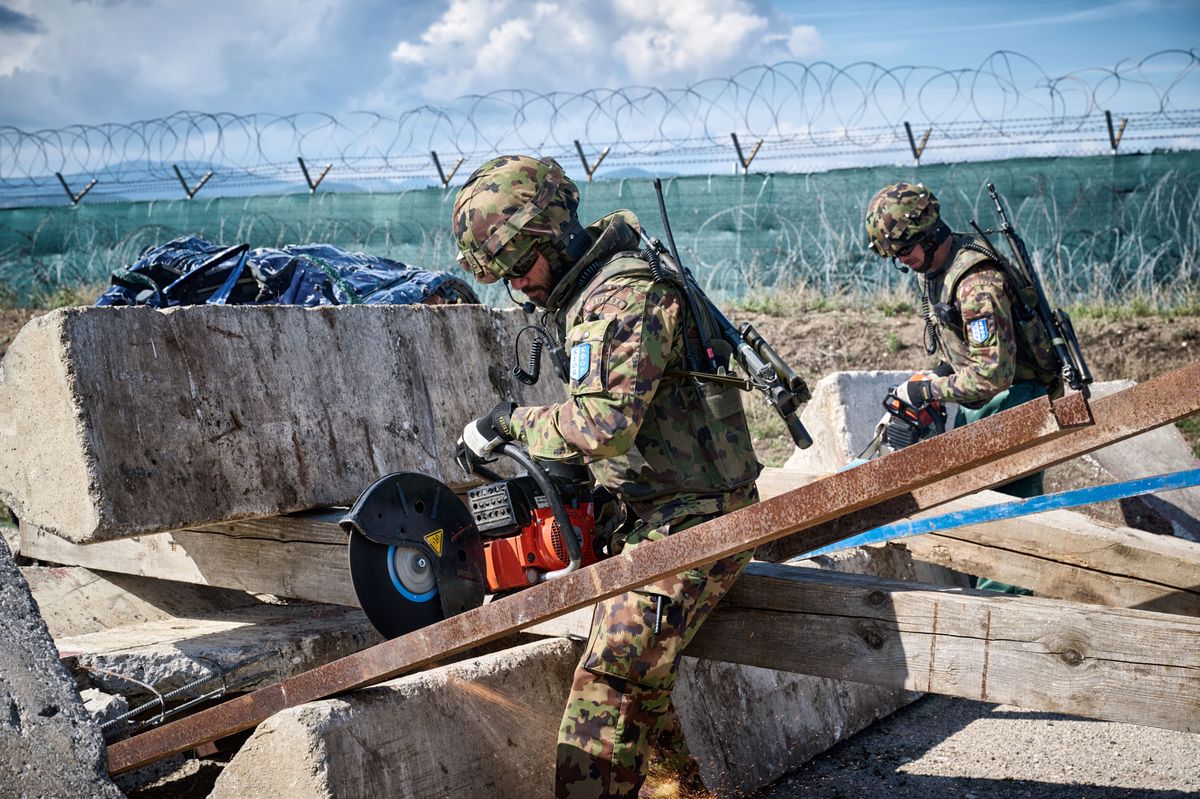 Deux soldats en uniforme militaire utilisent des outils pour couper des barres métalliques sur un site de travaux. Une clôture en fil barbelé est visible à l'arrière-plan.