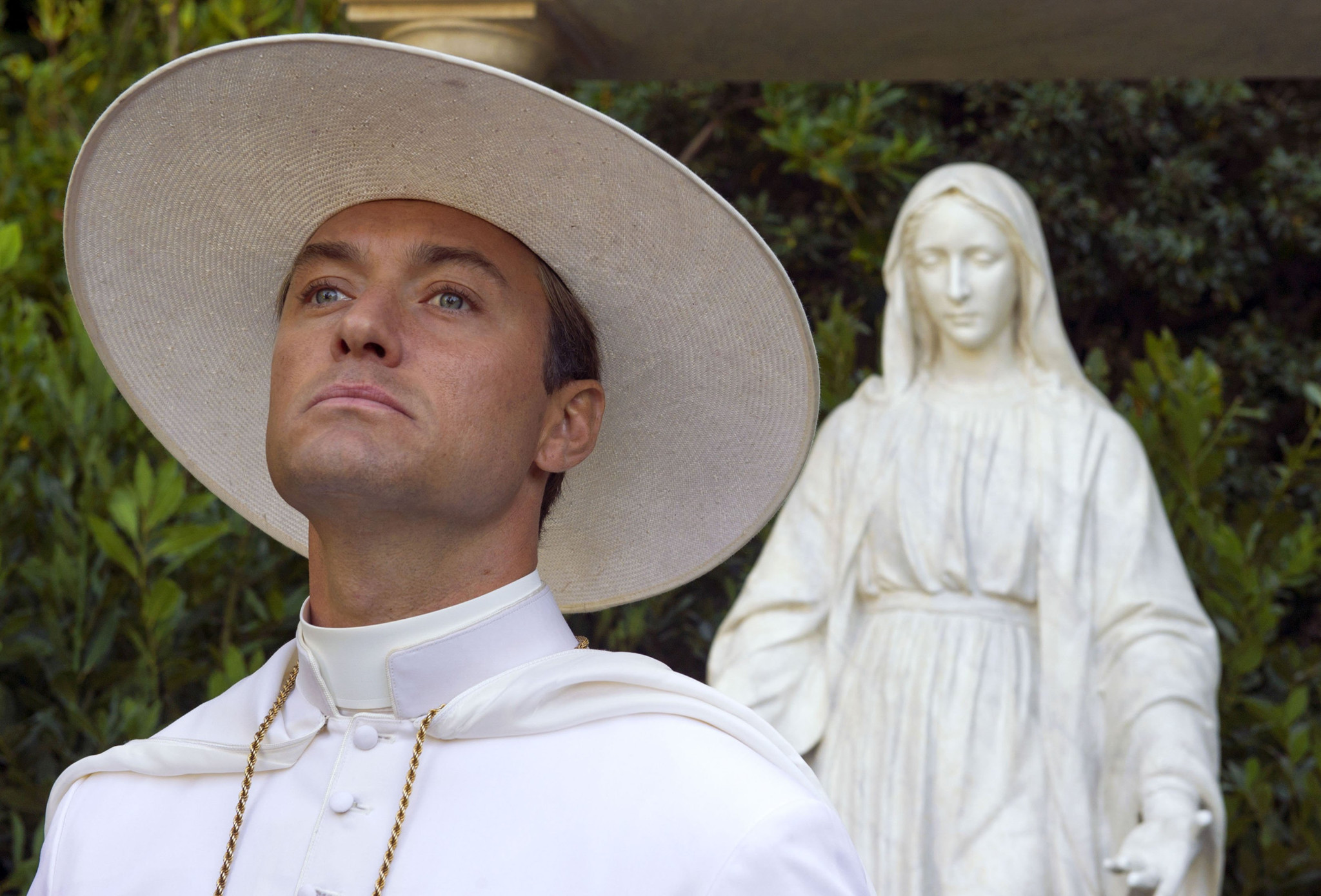 Jude Law dans le rôle du jeune pape, portant un chapeau papal blanc, avec une statue derrière lui, scène de la série ’The Young Pope’ de 2016.