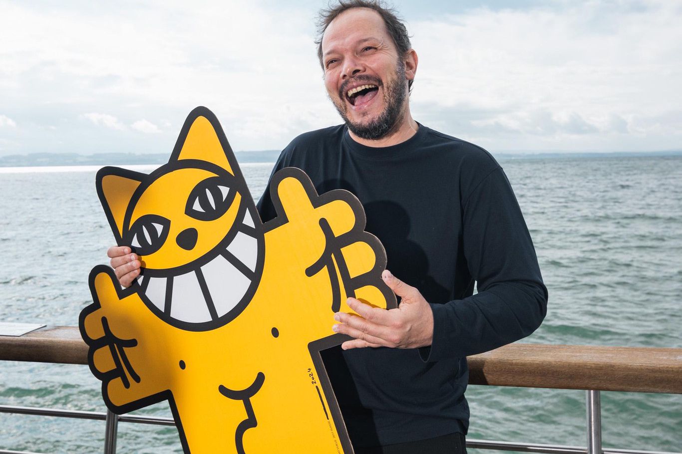 Homme souriant tenant une figure de chat jaune avec un large sourire, debout sur une promenade près de l'eau.
