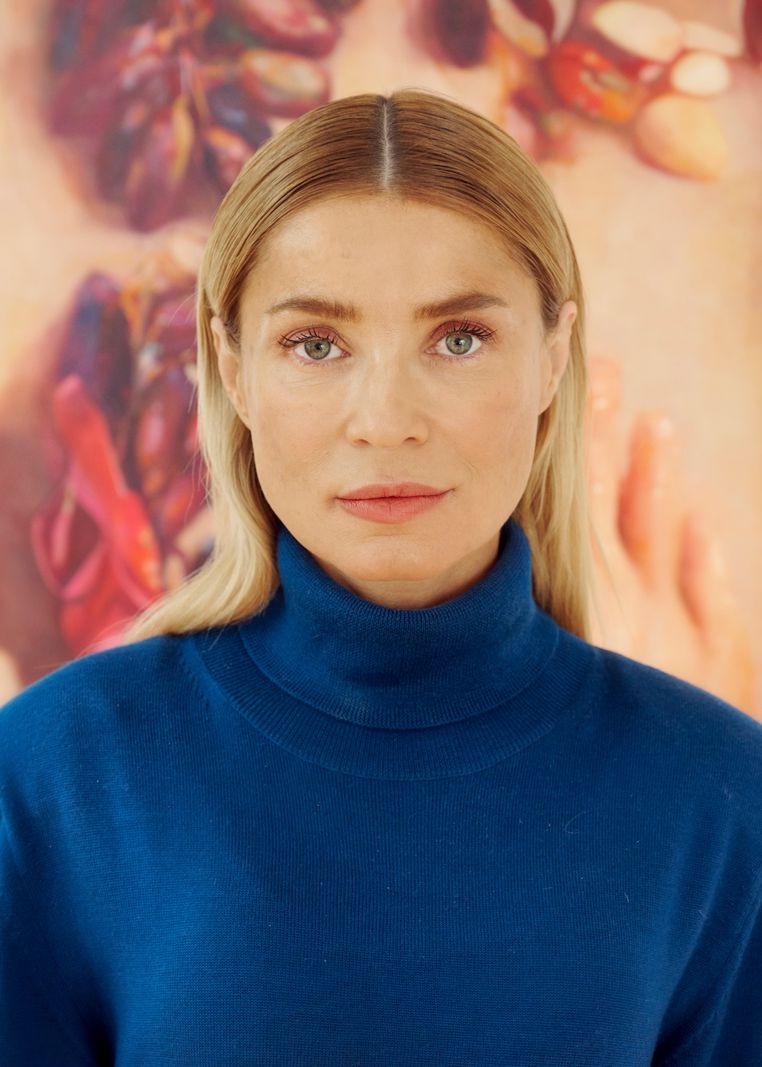 Une femme aux cheveux blonds et aux yeux clairs, portant un pull bleu à col roulé, se tient devant un arrière-plan coloré et flou représentant des objets organiques.