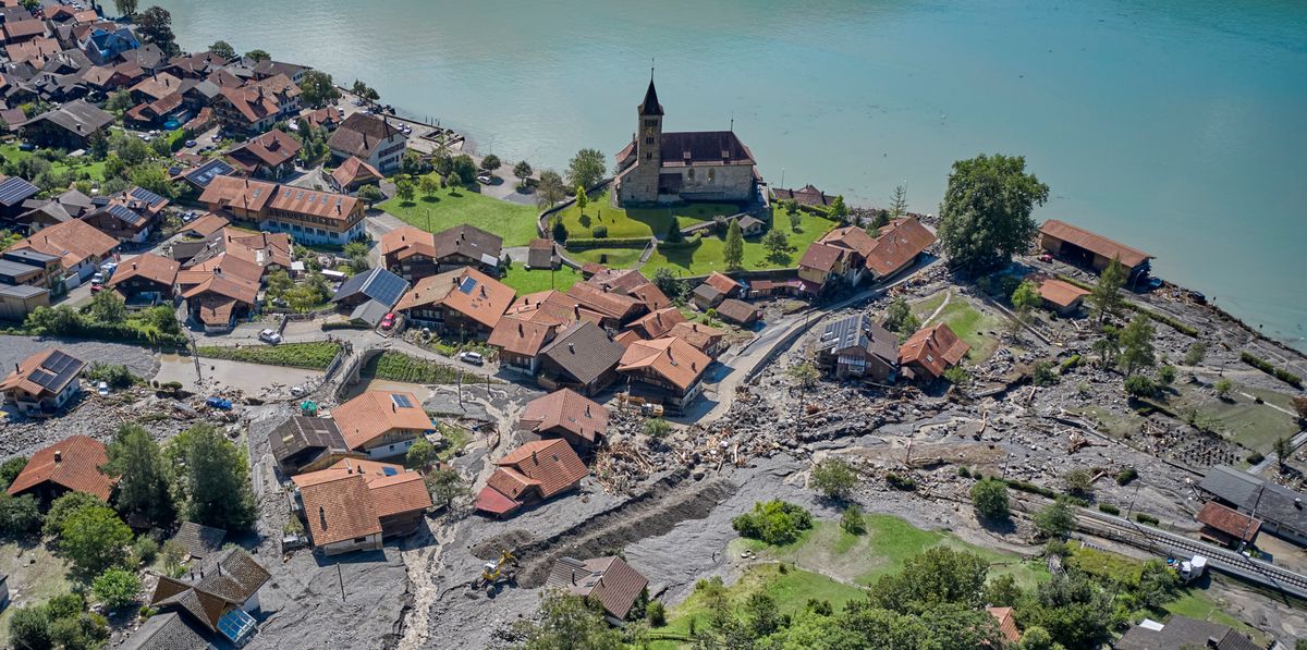 Luftaufnahme eines Dorfes am Seeufer, das von einer Überschwemmung betroffen ist. Schlamm und Trümmer bedecken Strassen und Grundstücke, während ein paar Häuser beschädigt erscheinen. Eine Kirche steht prominent im Dorf.