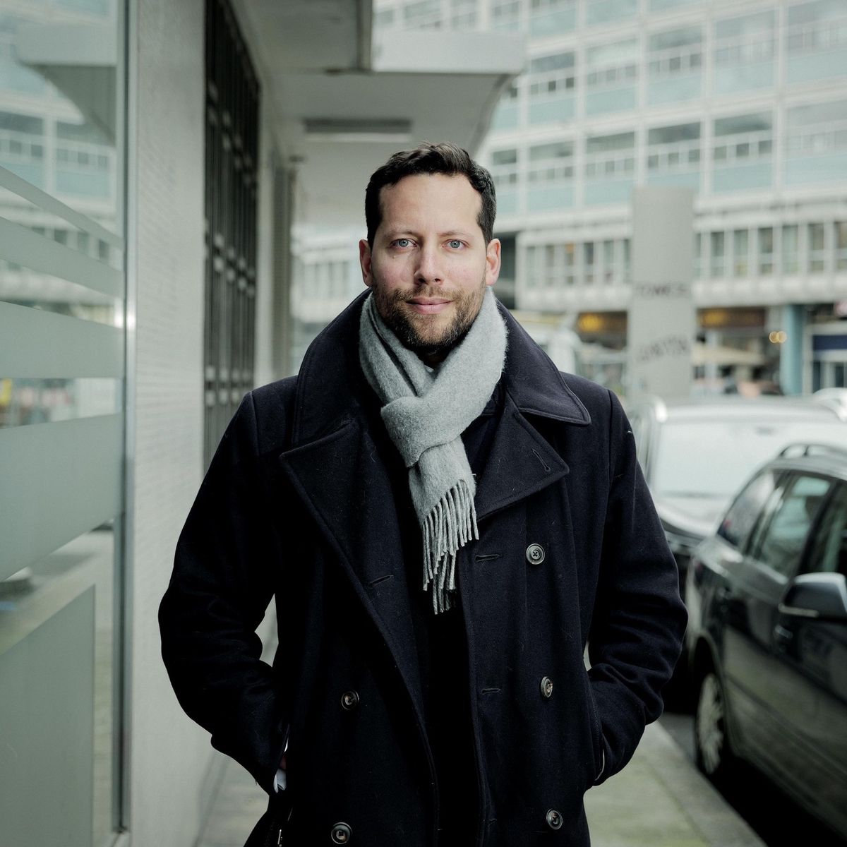 Un homme en manteau noir et écharpe grise se tient devant un bâtiment à Genève, discutant de l’initiative panneaux solaires.