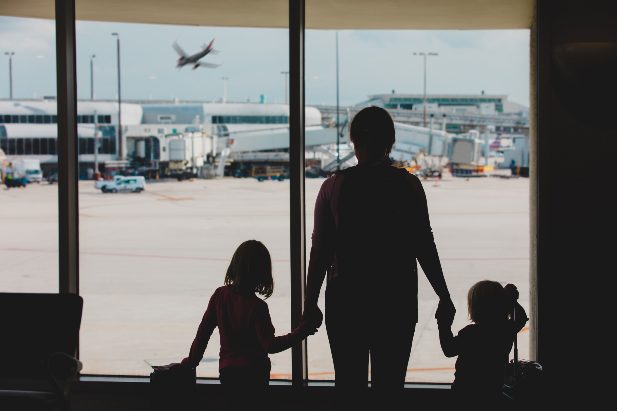 Silhouette einer Mutter mit zwei Kindern am Flughafenfenster, die ein startendes Flugzeug beobachten – Familienreise Silhouette einer Mutter mit zwei Kindern am Flughafenfenster, die ein startendes Flugzeug beobachten – Familienreise