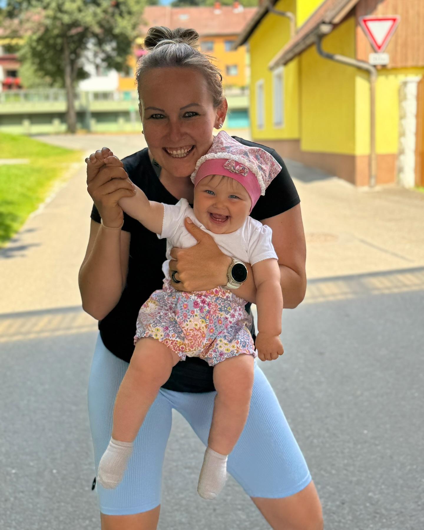 Frau hält ein lächelndes Baby im Freien, vor einem gelben Gebäude. Beide wirken fröhlich und geniessen die Sonne.