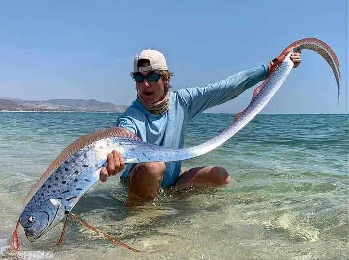 Un pêcheur agenouillé dans l’eau peu profonde tient fièrement un énorme poisson-ruban argenté aux nageoires rouges.