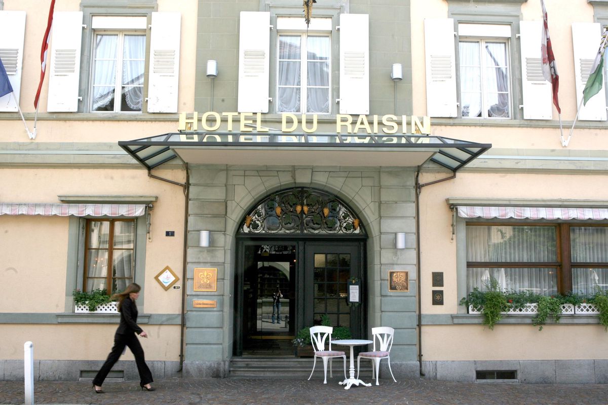 Façade de l’Hôtel du Raisin à Cully avec une femme passant devant l’entrée. Inscription ’Hôtel du Raisin’ visible.
