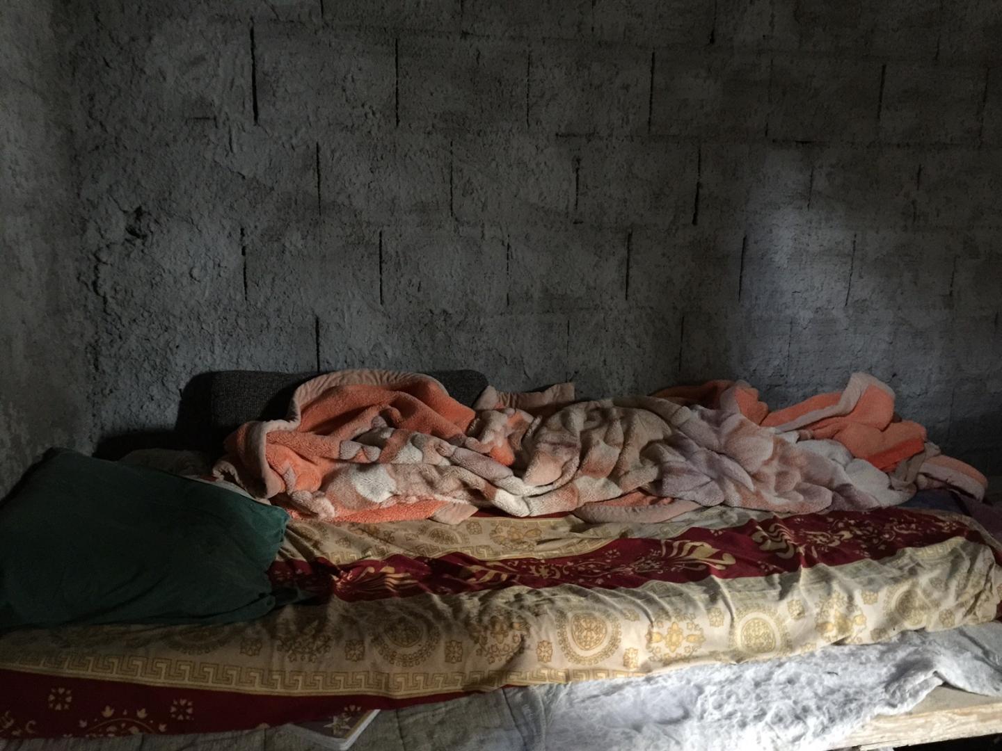 Un lit simple avec des couvertures colorées et un oreiller vert, placé contre un mur en béton gris dans une pièce sombre.