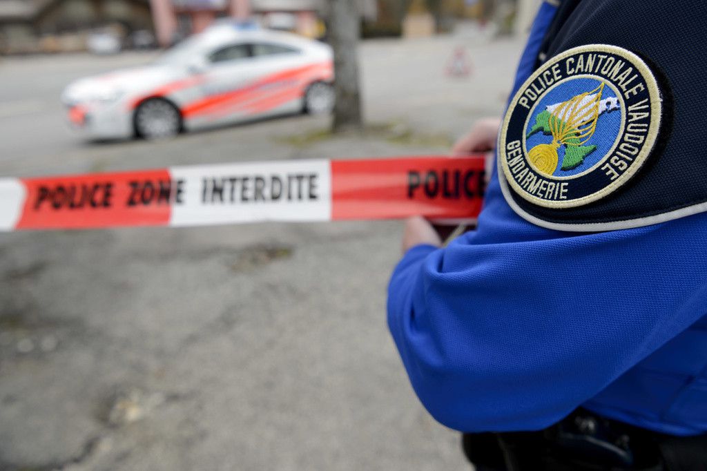 Un policier de la gendarmerie cantonale tient un ruban de sécurité marqué 'POLICE ZONE INTERDITE', avec une voiture de police en arrière-plan.
