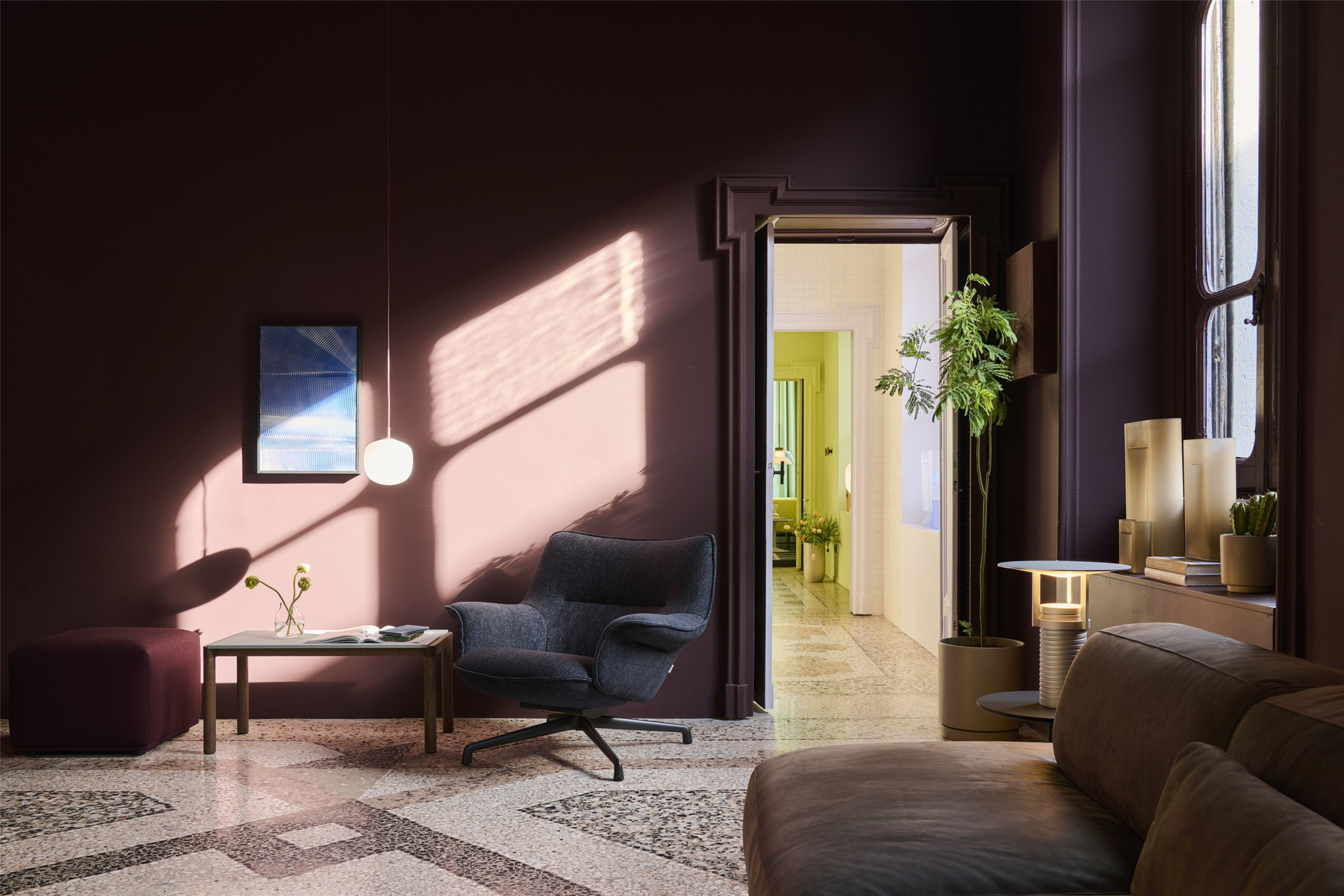 Elegantes Wohnzimmer mit dunkelroten Wänden, grauem Sessel, Terrazzoboden und Blick durch eine Türöffnung in einen hellen Flur.