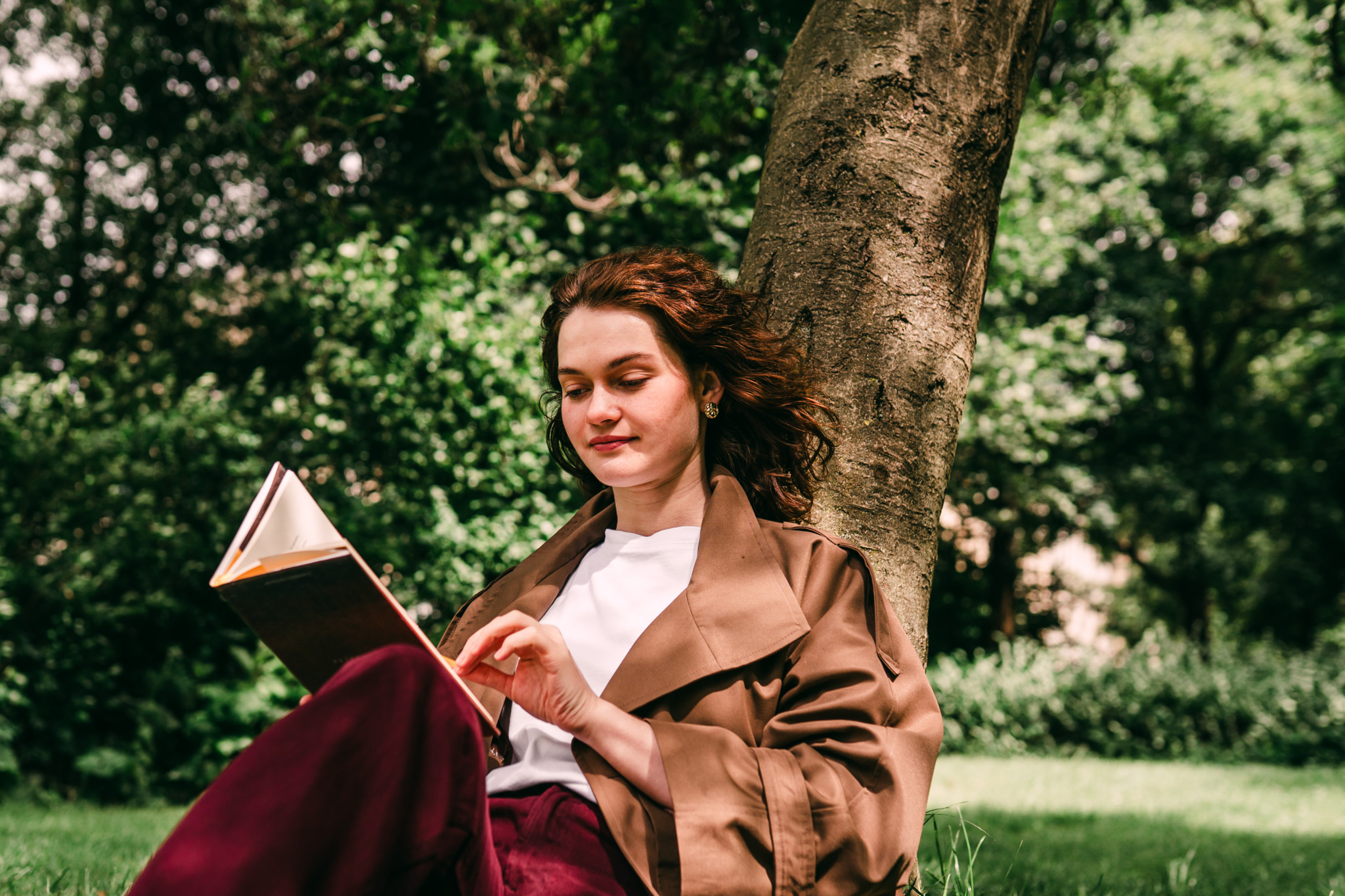 Junge Frau mit lockigem rotbraunem Haar liest lächelnd ein Buch, an einen Baumstamm gelehnt, im Park – Trenchcoat und burgunderrote Hose.
