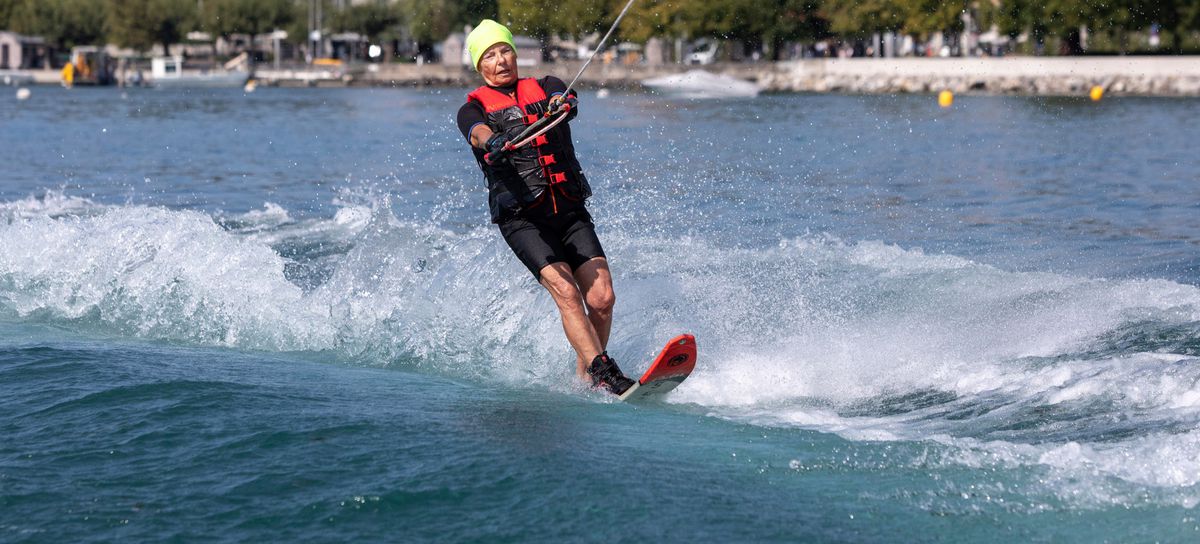 Personne pratiquant le ski nautique sur un lac avec des bâtiments historiques et des arbres en arrière-plan.