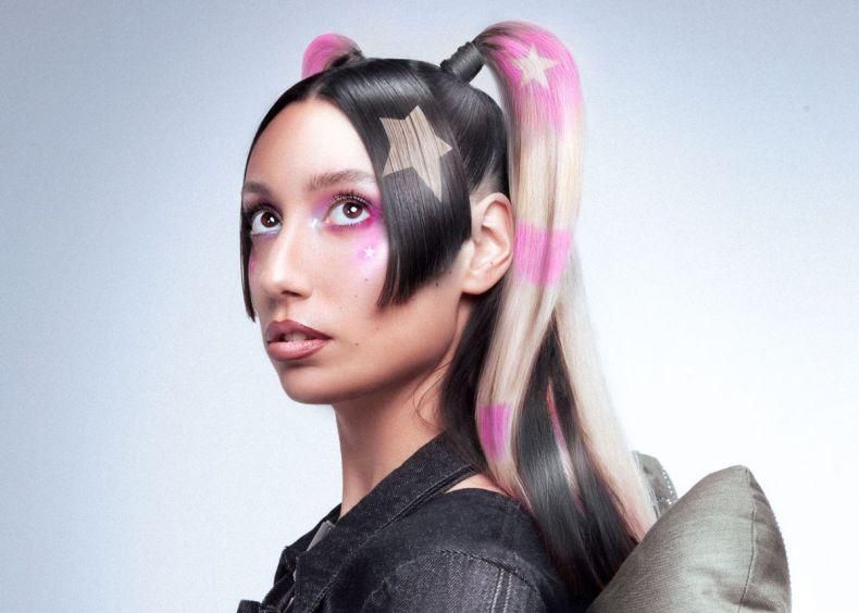 Une femme avec des cheveux coiffés en doubles couettes roses et noires, ornées d’étoiles, regarde vers le haut.