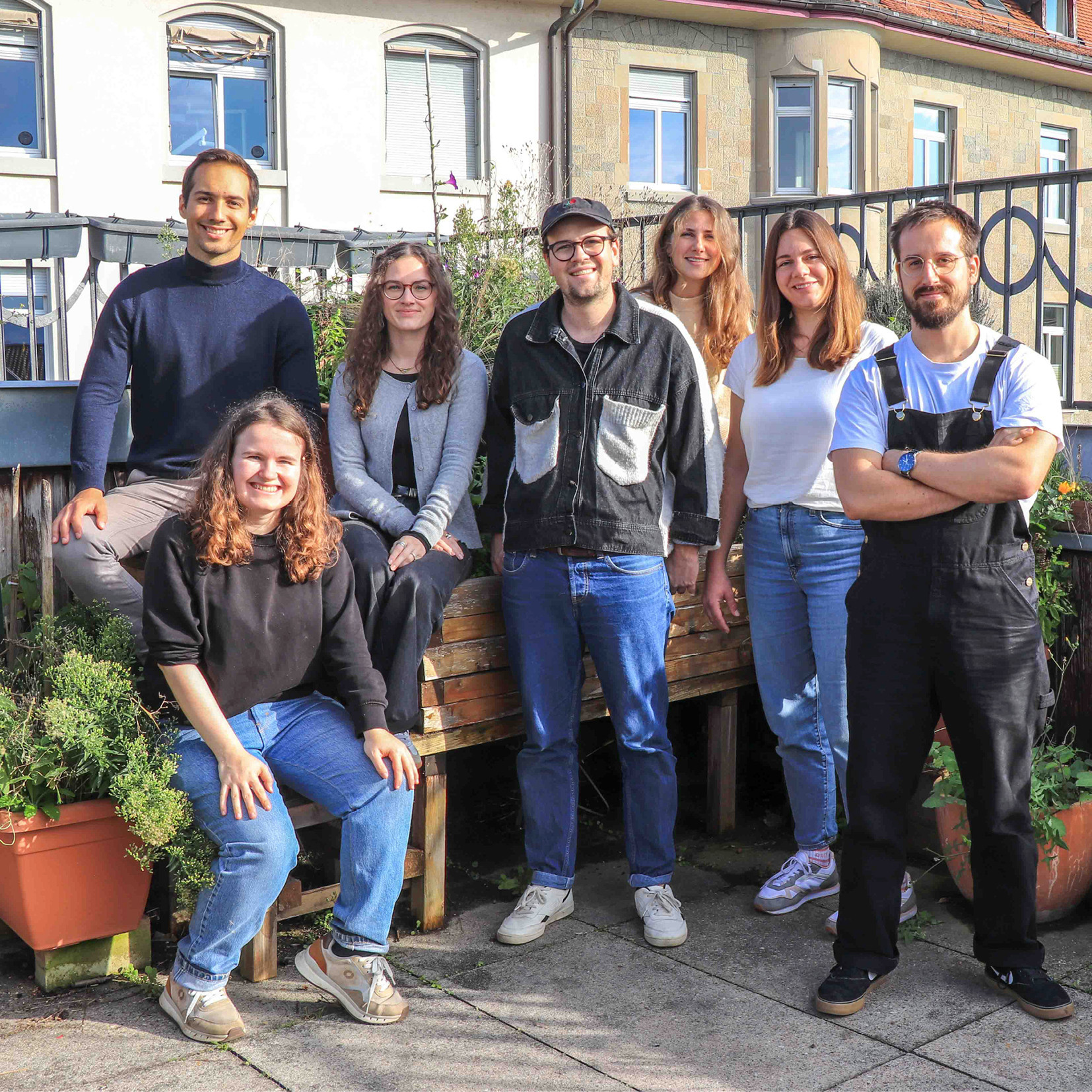 Eine Gruppe von sieben jungen Erwachsenen posiert lächelnd auf einer Terrasse mit Pflanzen im Hintergrund.