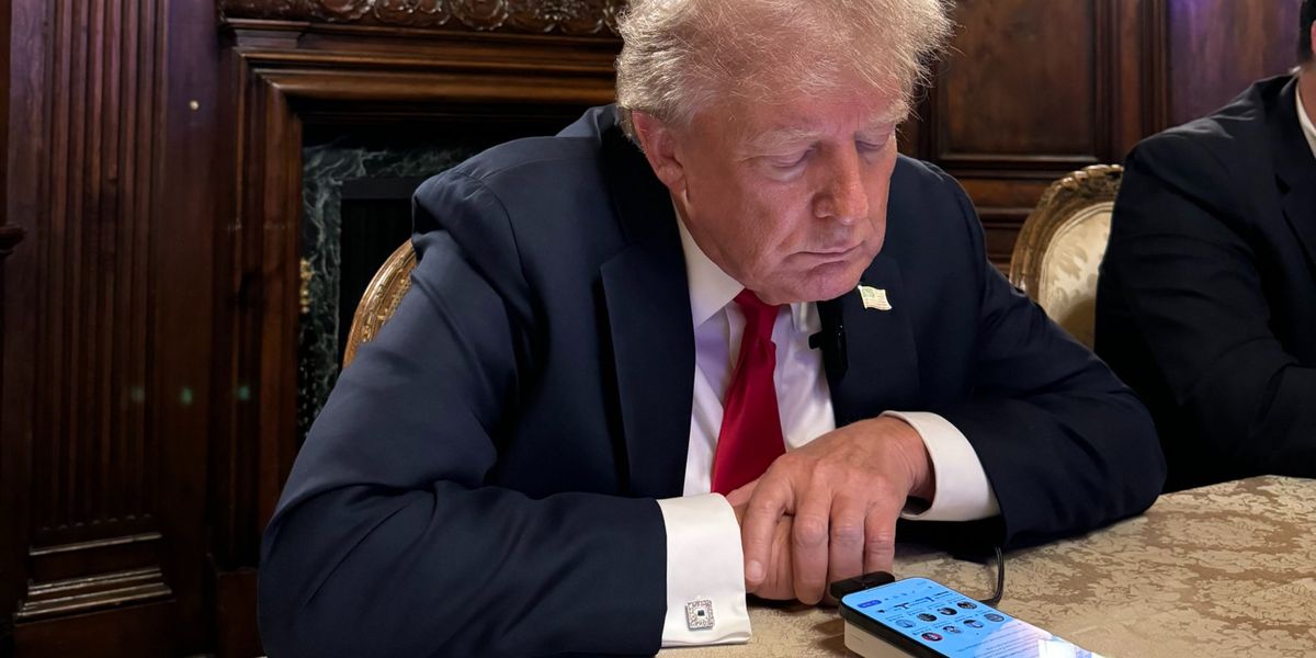 Ein älterer Mann in einem Anzug sitzt an einem Tisch und blickt konzentriert auf ein Mobiltelefon. Hinter ihm befindet sich ein reich verziertes Holzkabinett mit einem Gemälde von Segelschiffen.