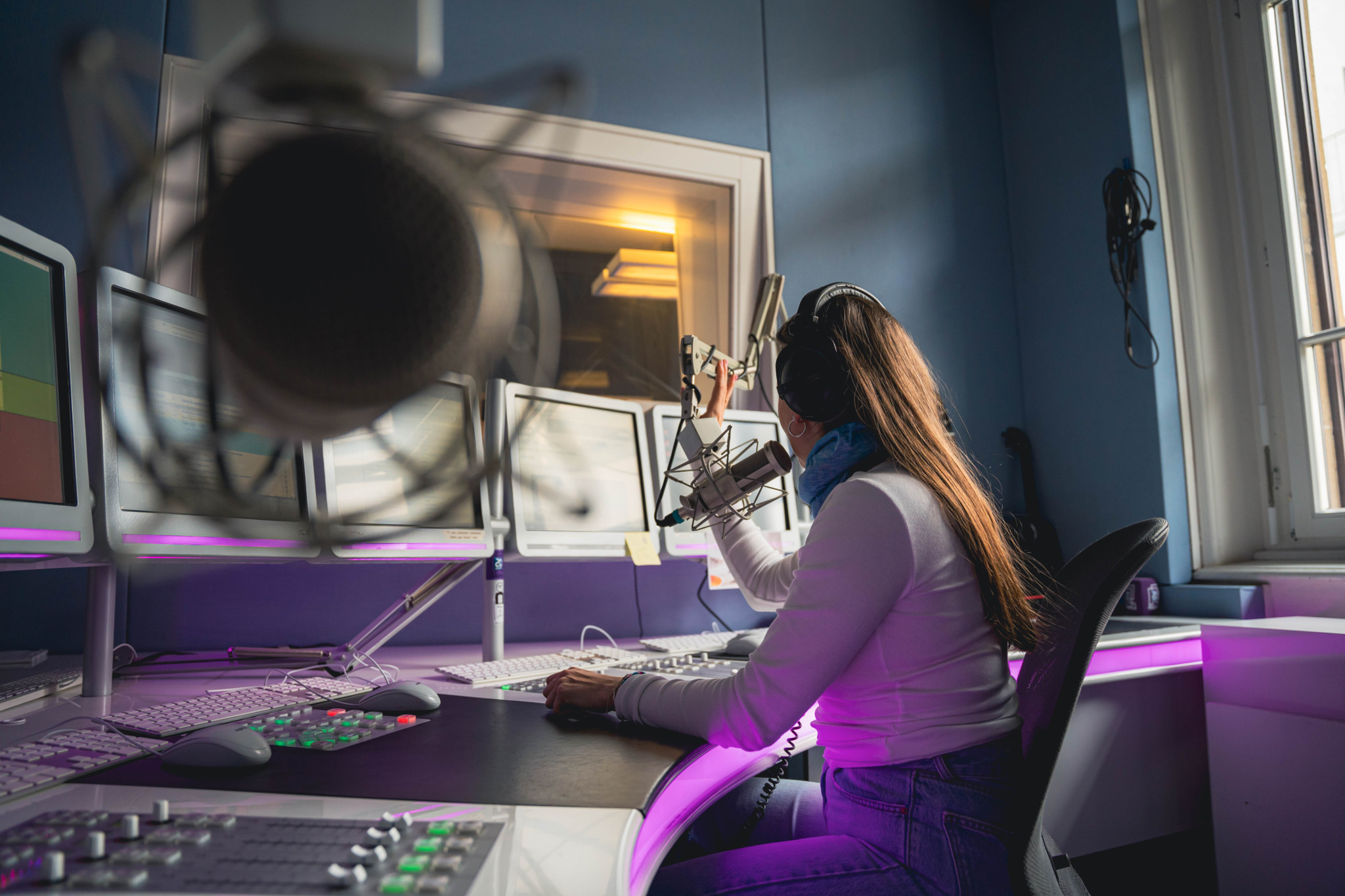 Radiomoderatorin in einem Studio spricht in ein Mikrofon, umgeben von Bildschirmen und Mischpult.