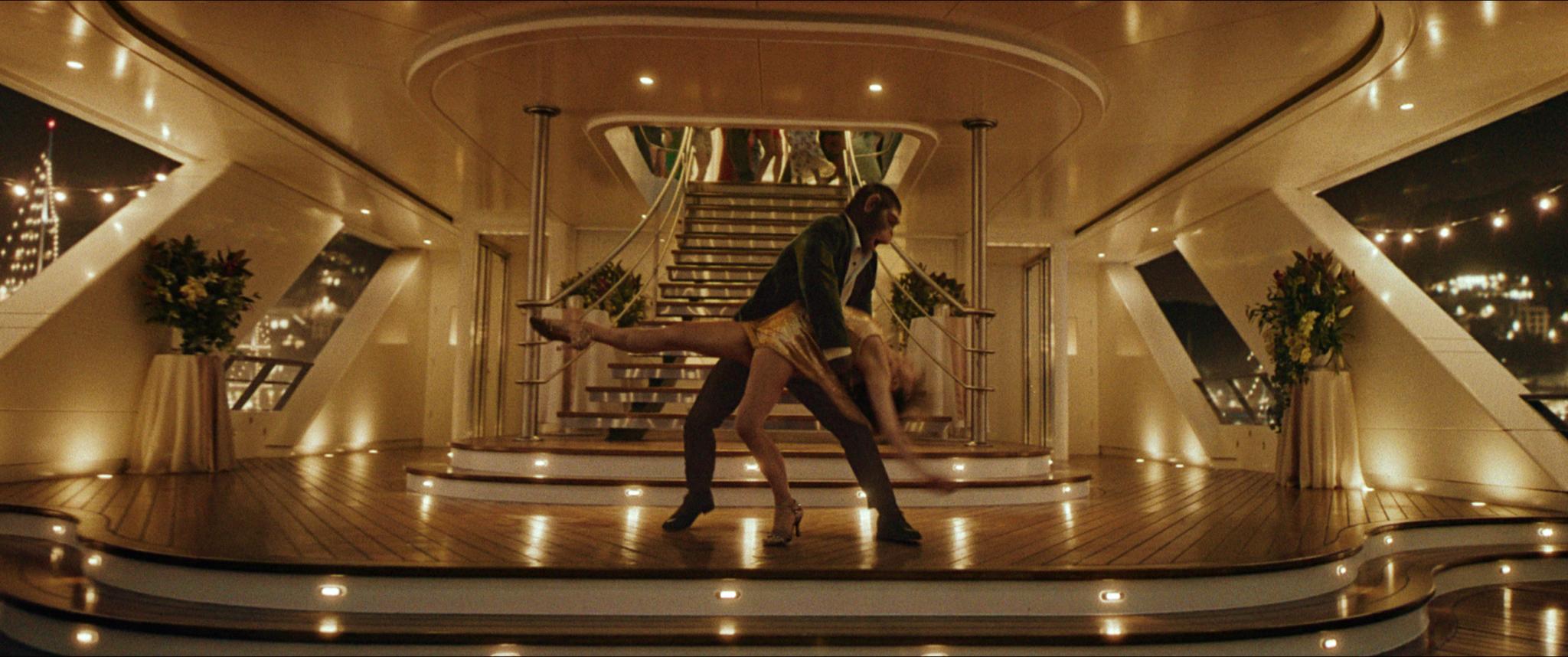 Danseurs exécutant une pose sur le pont luxueux d’un yacht éclairé, avec un escalier central élégant en arrière-plan.