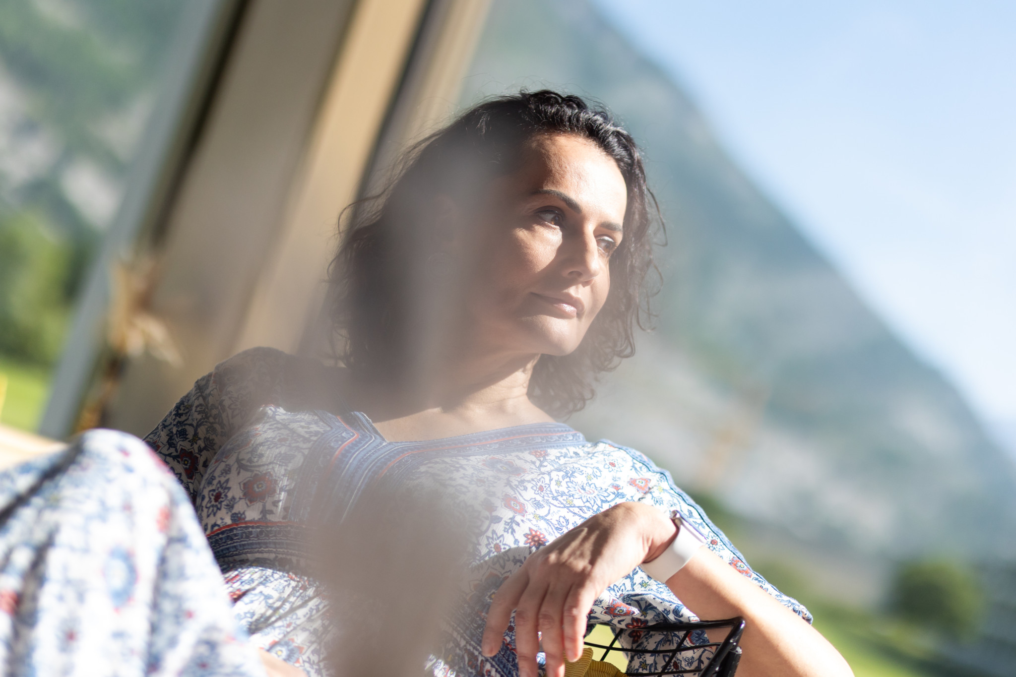 Amina Ovcina Cajacob sitzt nachdenklich vor einem Fenster in Chur, mit Berglandschaft im Hintergrund.