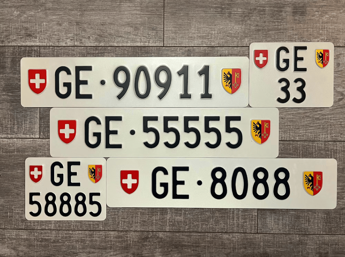Plaques d’immatriculation suisses avec blason de Genève, montrant différents numéros comme 90911 et 58885, alignées sur un sol en bois.