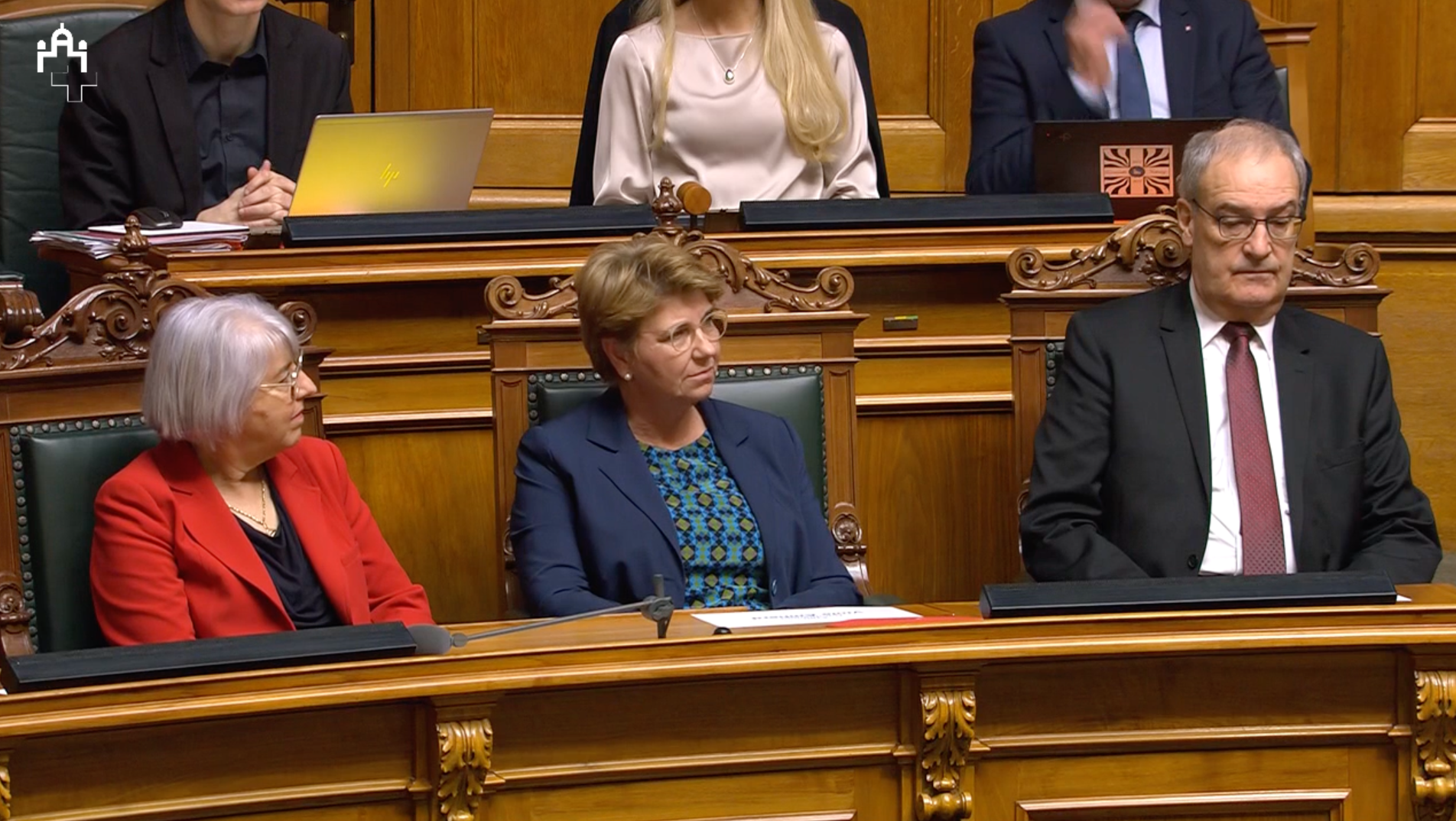 Trois personnes assises dans une salle de conférence en bois, écoutant attentivement.