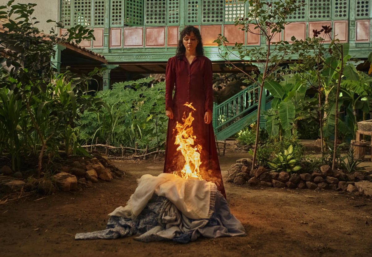 Loren Sofía interprète Amaranta, debout devant un feu de linge dans un jardin luxuriant, scène de ’Cien Años de Soledad’.