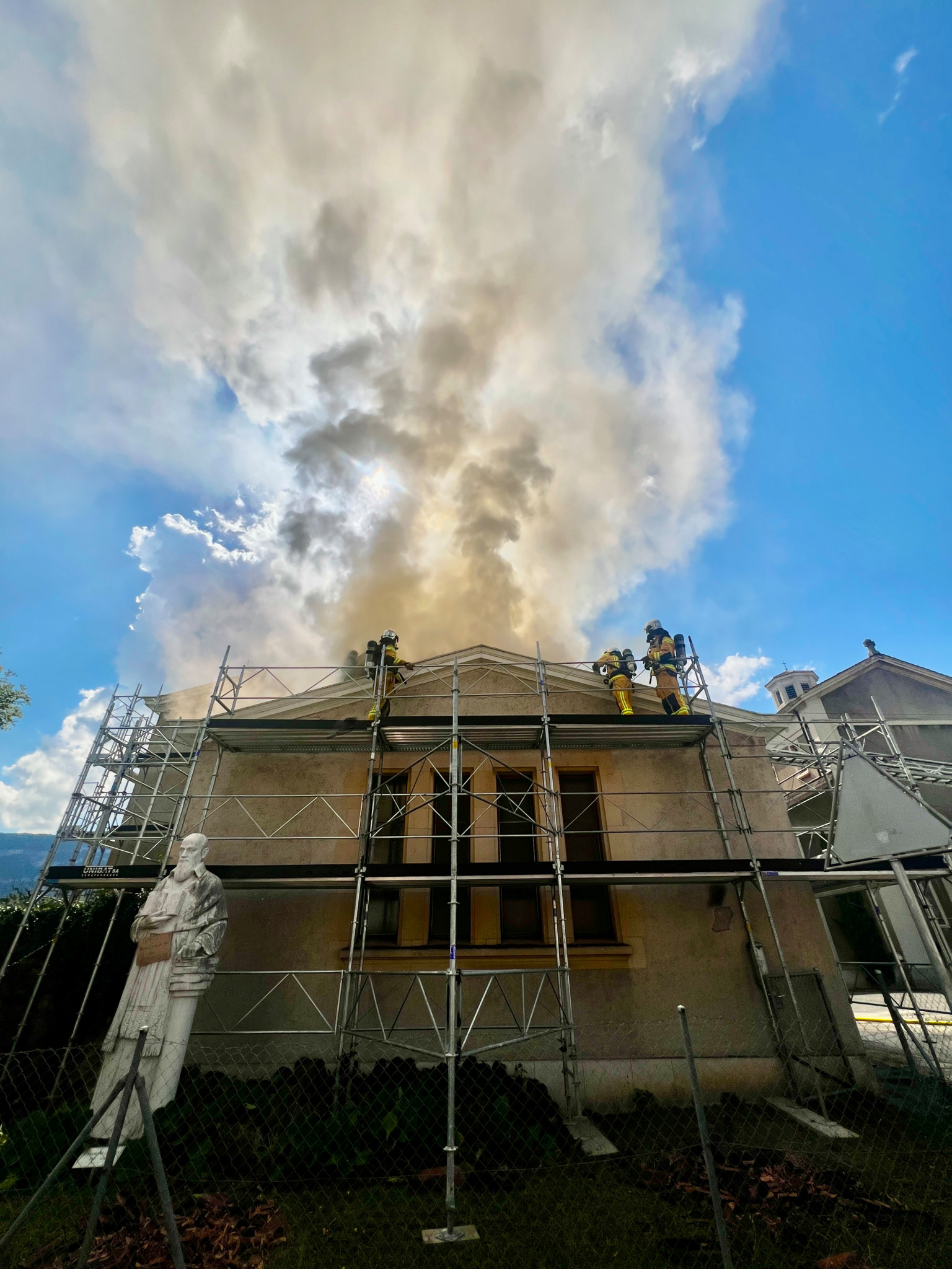 Des pompiers interviennent sur le toit d'un bâtiment en feu, avec une statue de marbre blanche au premier plan et des échafaudages entourant la structure, sous un ciel bleu avec une colonne de fumée épaisse.