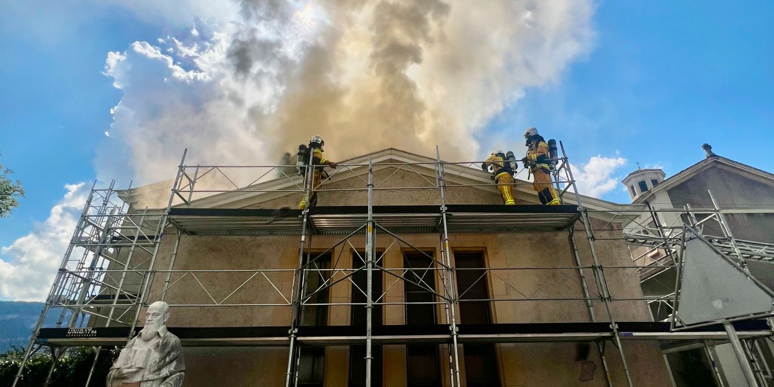 Des pompiers interviennent sur le toit d'un bâtiment en feu, avec une statue de marbre blanche au premier plan et des échafaudages entourant la structure, sous un ciel bleu avec une colonne de fumée épaisse.