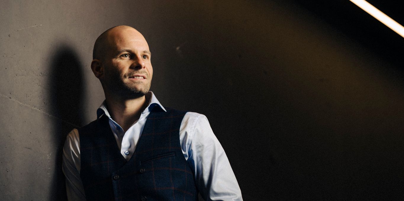 Nicolas Fluri, nouveau directeur des arbitres suisses de hockey sur glace, souriant et posant contre un mur sombre à la Vaudoise Arena de Prilly-Malley, le 26 septembre 2024.