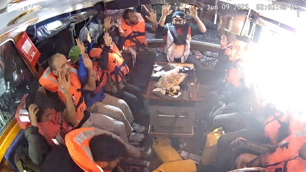Menschen in einem Boot tragen Sicherheitswesten und halten ihre Hände hoch, als ob sie sich schützen oder gestikulieren. Ein Mann trägt einen blauen Helm. Ein Tisch mit einer Karte und anderen Gegenständen steht in der Mitte.